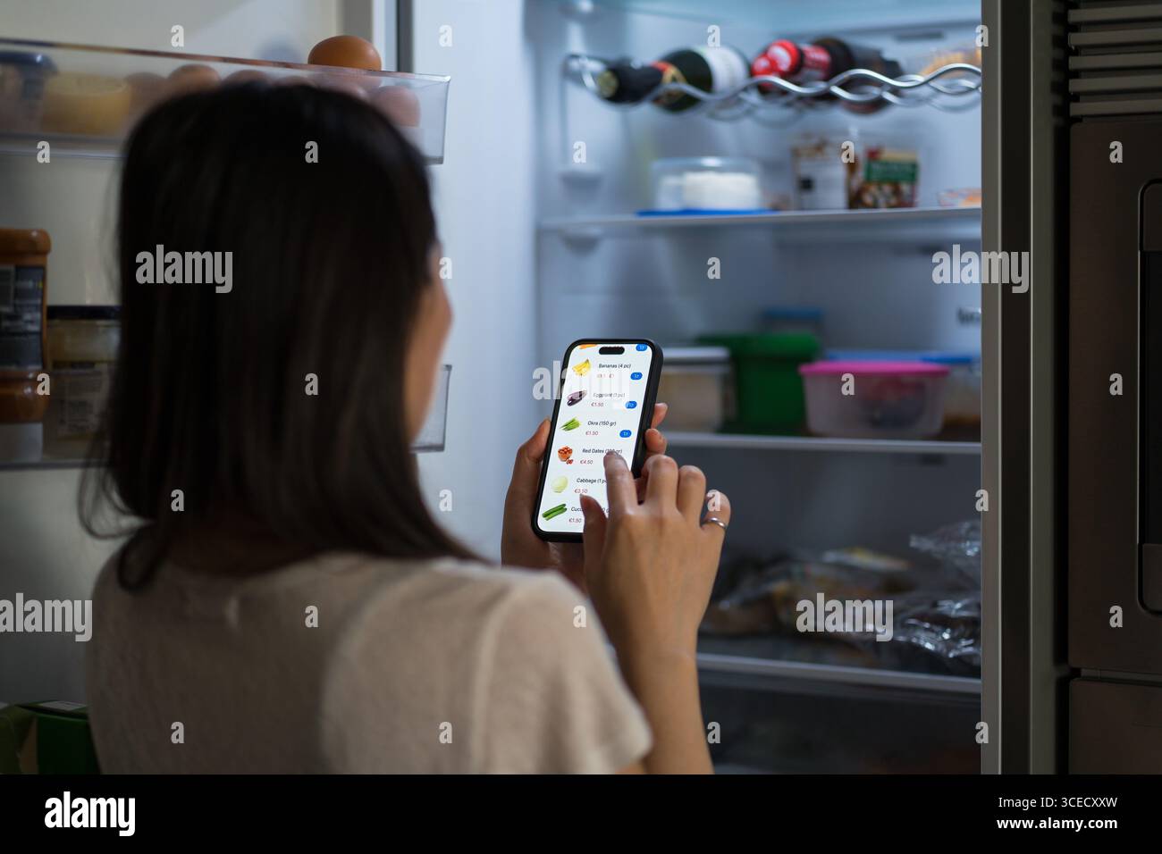 Vista posteriore di una donna irriconoscibile che usa lo smartphone per fare acquisti online di fronte a un frigorifero aperto. Esplora opzioni salutari tramite un'app, in Foto Stock