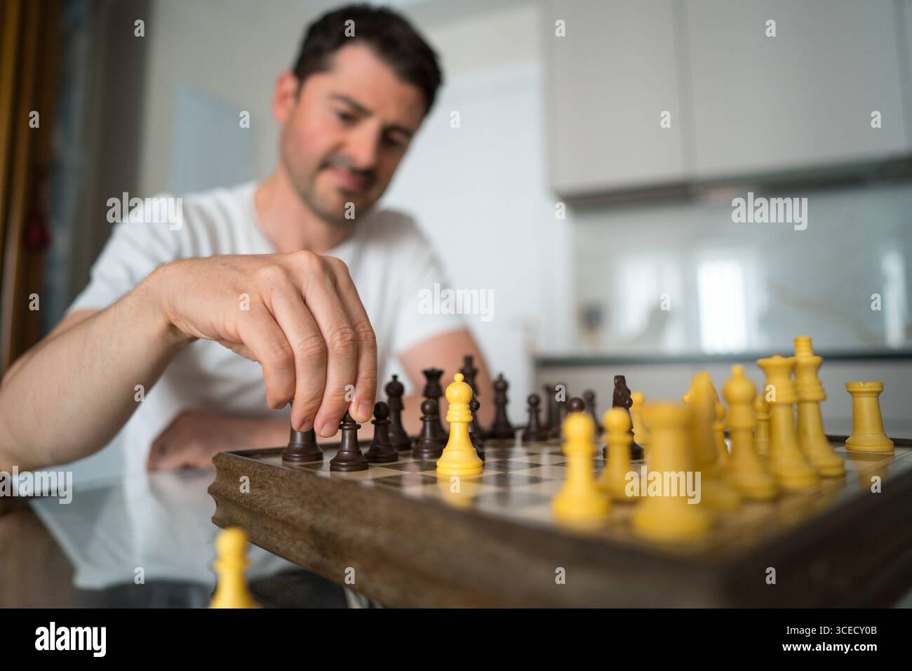 Un uomo maturo siede a un tavolo, profondamente impegnato in una partita a scacchi a casa. Considera con attenzione la sua prossima mossa, creando un momento di calma concentrazione Foto Stock