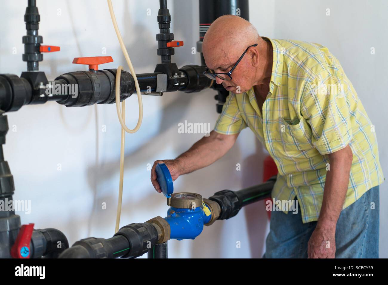 Un anziano ispeziona attentamente un sistema di contatori dell'acqua in un ambiente rurale. Indossa una camicia e degli occhiali a quadri gialli, dimostrando cura ed esasperazione Foto Stock