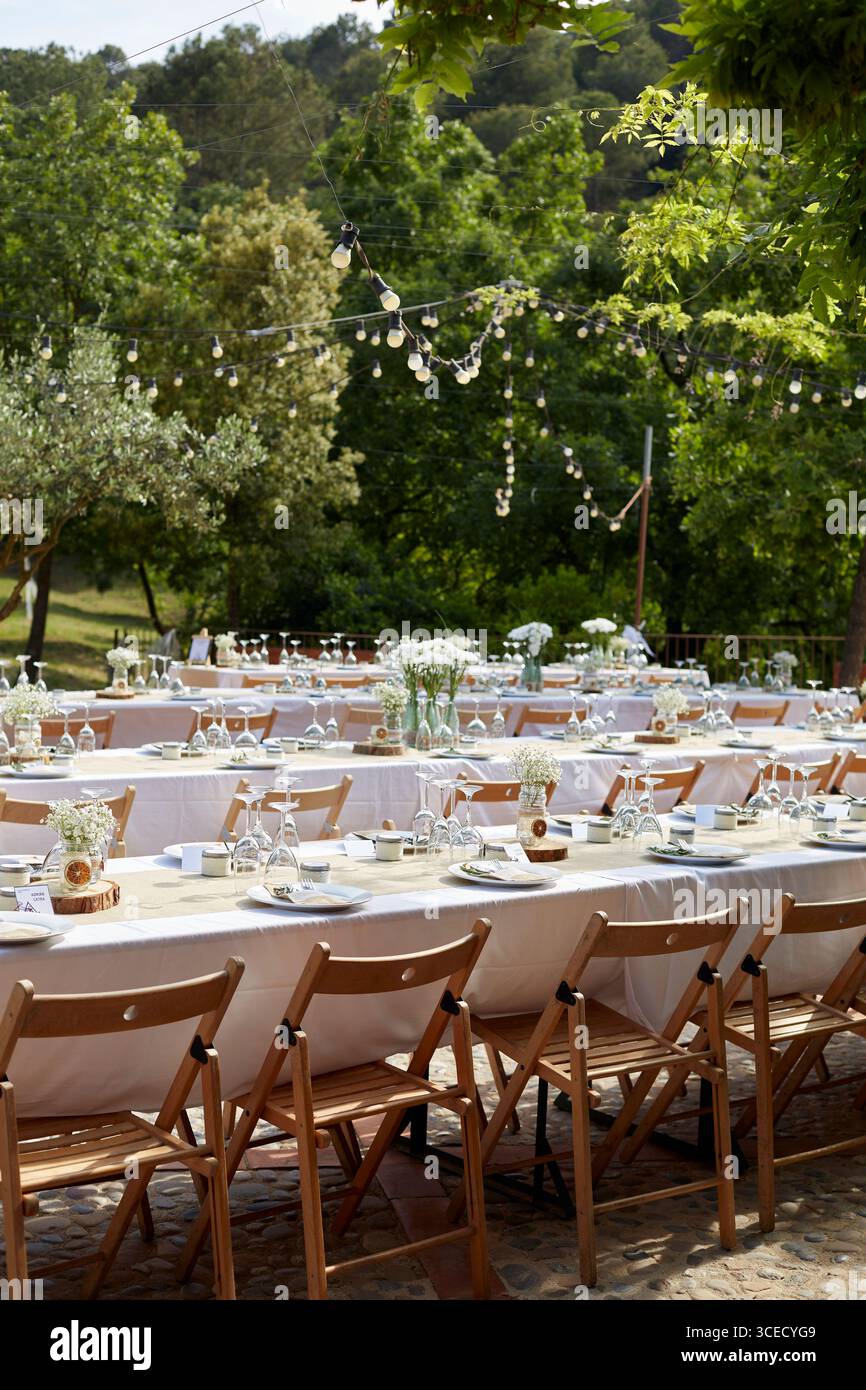 Elegante allestimento per matrimoni all'aperto con sedie e tavoli in legno, adornato con tovaglie bianche e vasi in vetro che reggono delicate composizioni floreali, unde Foto Stock