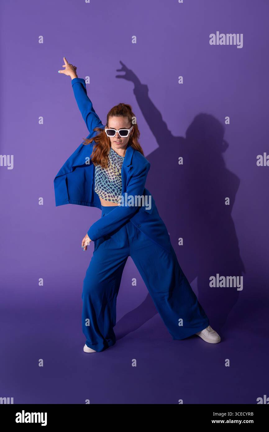 Una giovane donna con un vestito vivace danzerà energicamente su uno sfondo viola. La sua posa dinamica e il suo movimento espressivo catturano l'essenza del co Foto Stock