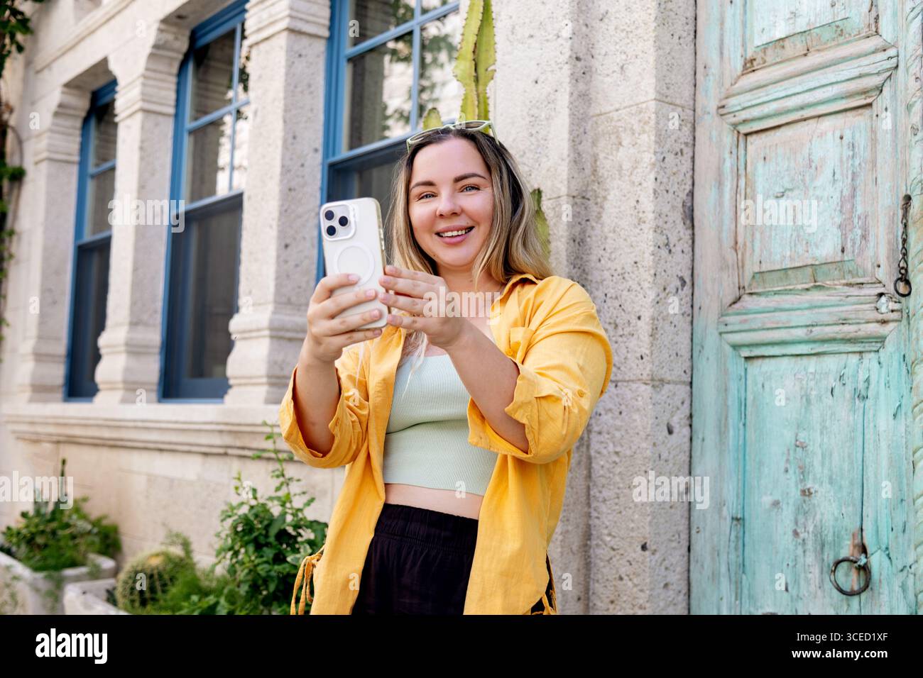 Una allegra turista che indossa una camicia gialla utilizza il suo smartphone per immortalare momenti indimenticabili durante il viaggio, davanti a una storica b Foto Stock