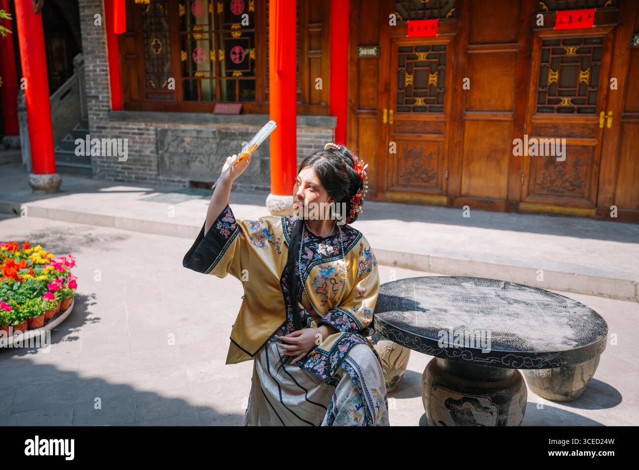 Una giovane donna con un abbigliamento tradizionale della dinastia Qing si siede graziosamente all'aperto a Pingyao, in Cina, mentre si ombreggia con un elegante ventilatore sotto la zampa Foto Stock