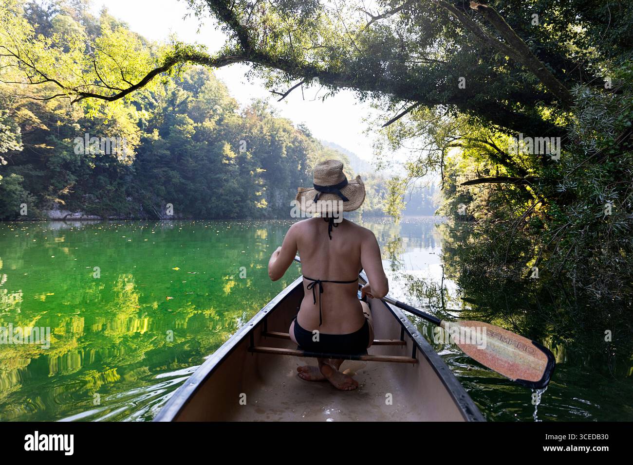 Vista posteriore di una turista in bikini che rema una canoa attraverso la panoramica gola del fiume Idrijca circondata da scogliere e alberi, Slovenia. Foto Stock