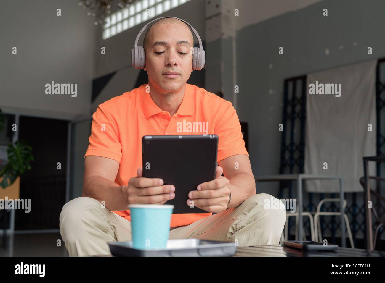 Bell'uomo di mezza età con una polo arancione al tavolino da caffè con una tazza di caffè, cuffie e tablet digitali. Tecnologia e cas Foto Stock