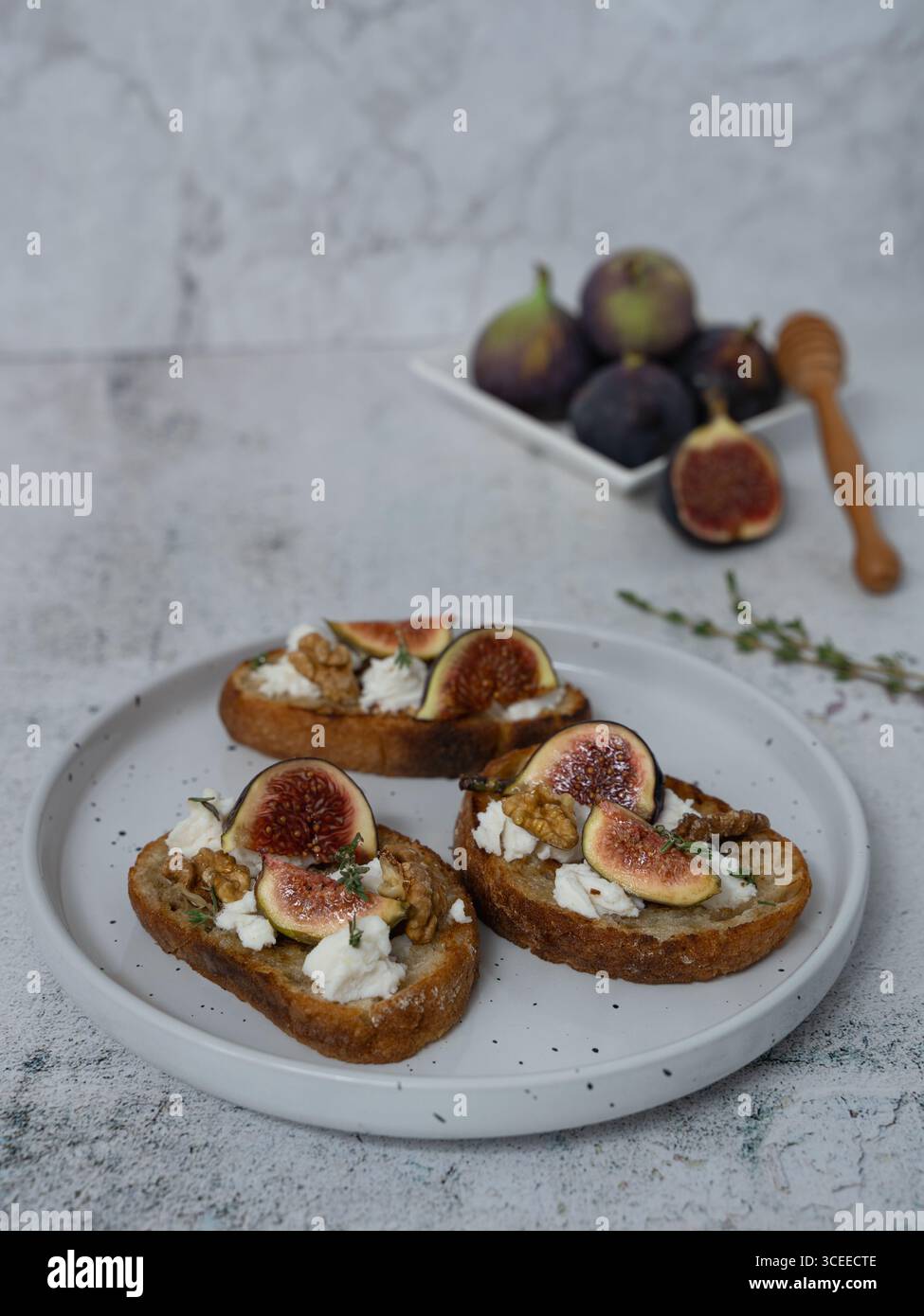 Primo piano verticale vista frontale di tre toast di fichi e capra su piatto bianco con timo, forchetta, sfondo grigio, fichi su piatto separato, toast in b Foto Stock