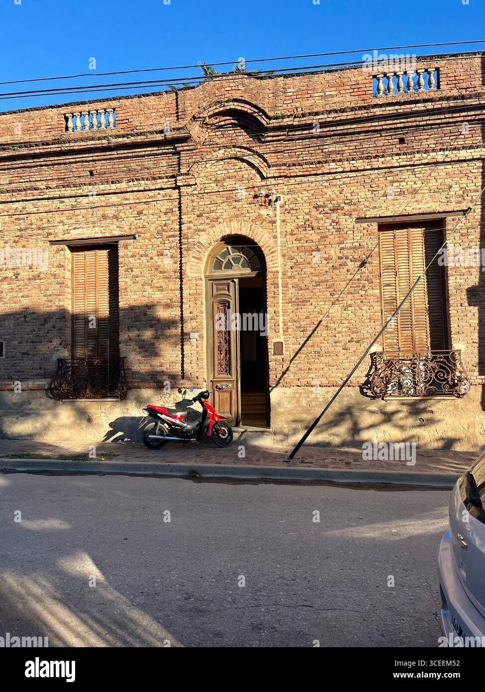 Una rustica casa in mattoni con porta ad arco e persiane in legno, con una motocicletta parcheggiata davanti su una strada soleggiata. - Immagine stock catturata con smartphone