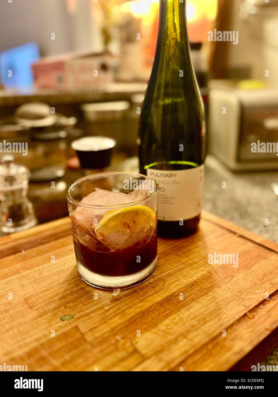 Un cocktail scuro servito in un bicchiere di roccia con ghiaccio e un tocco di limone su una tavola da bar in legno, con una bottiglia e l'attrezzatura da bar sullo sfondo. - Immagine stock catturata con smartphone