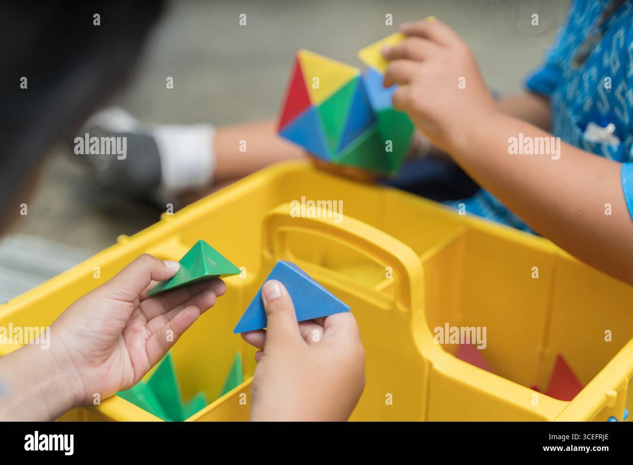 Bambini che utilizzano blocchi geometrici colorati da una scatola gialla per giocare e imparare, simboleggiando l'istruzione, la creatività, il divertimento, lo sviluppo, immaginazione, abilità Foto Stock