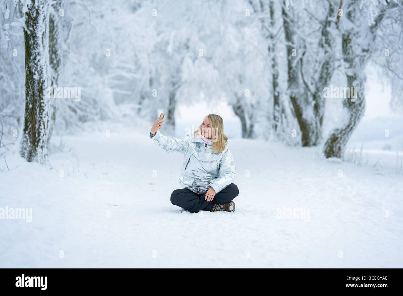 Una donna seduta su un sentiero innevato cattura selfie in mezzo a una foresta innevata. Vestita con calore con cappotto invernale, trasuda gioia e serenità nel Foto Stock