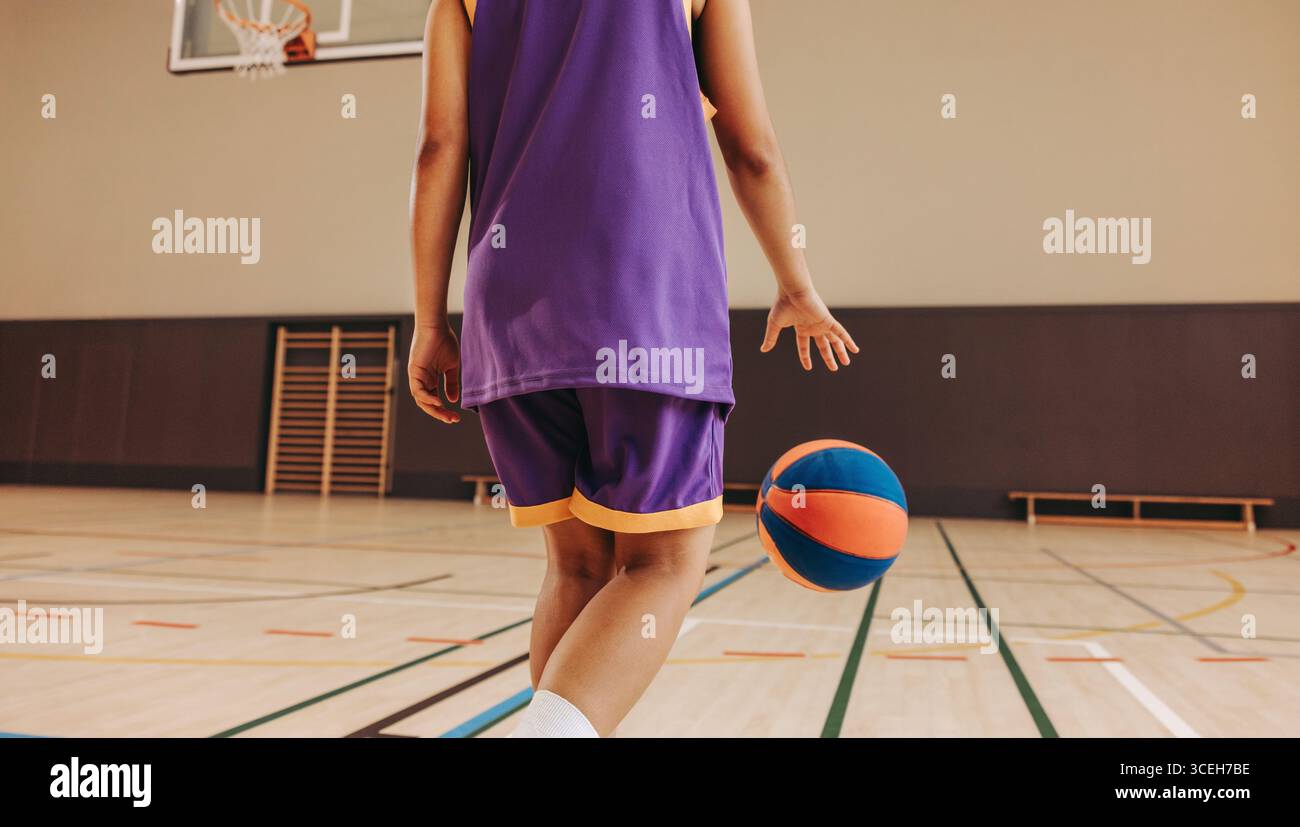 Un giovane giocatore di abbigliamento sportivo viola drizza un pallacanestro su un campo di legno al chiuso, dando prova di atleticità. L'immagine trasmette energia, messa a fuoco e crogiolo Foto Stock