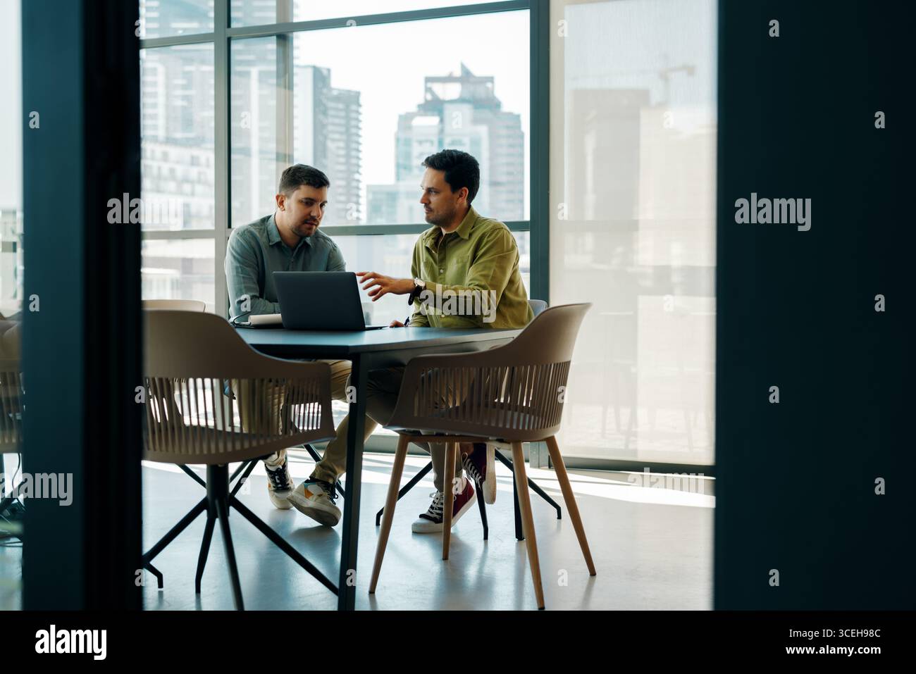 Due giovani che lavorano insieme a un progetto, seduti a un tavolo con un portatile. L'ambiente è uno spazio open Office con finestre in vetro e vista su un Foto Stock