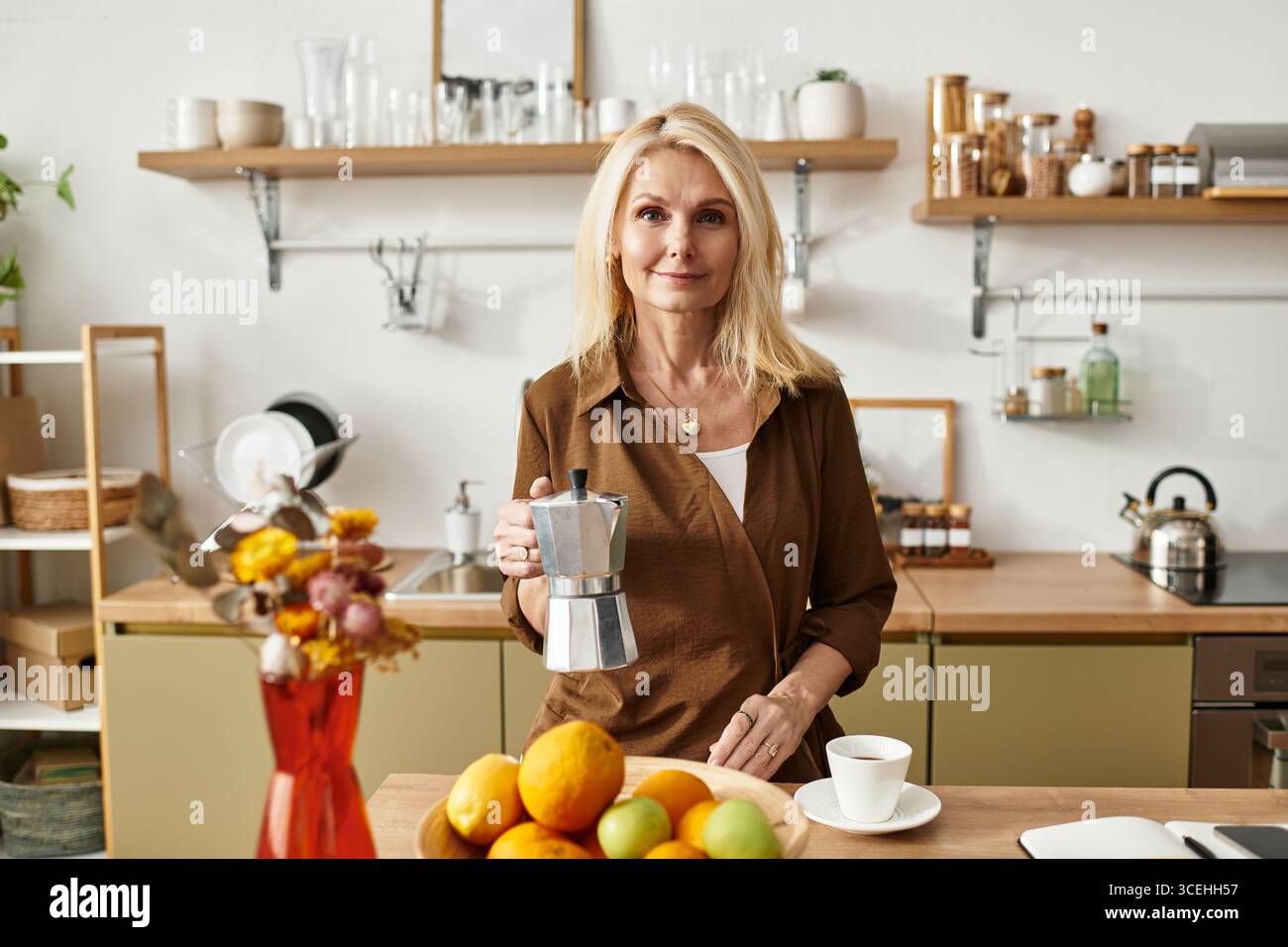 Una bella donna anziana si trova in una cucina moderna con una macchina per il caffè e frutta fresca. Foto Stock