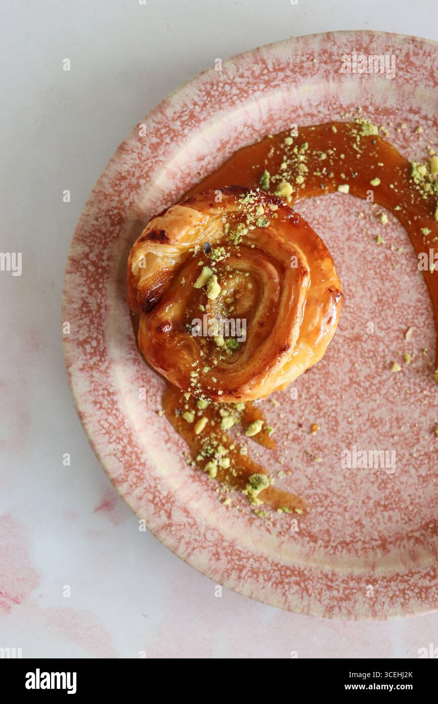 Una vista dall'alto verso il basso di un rotolo di pasta sfoglia gourmet, condito con una glassa dolce al caramello e cosparso di pistacchi tritati su un piatto rosa rustico. Foto Stock