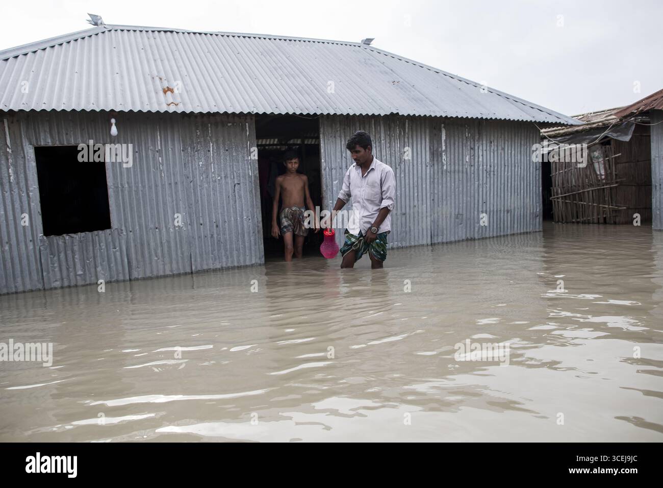 Kurigram, Bangladesh - 21 giugno 2022: Vista di una casa in metallo ondulato parzialmente sommersa in acque alluvionali, con un uomo e un bambino che attraversano le profondità torbide vicino alla porta. Foto Stock