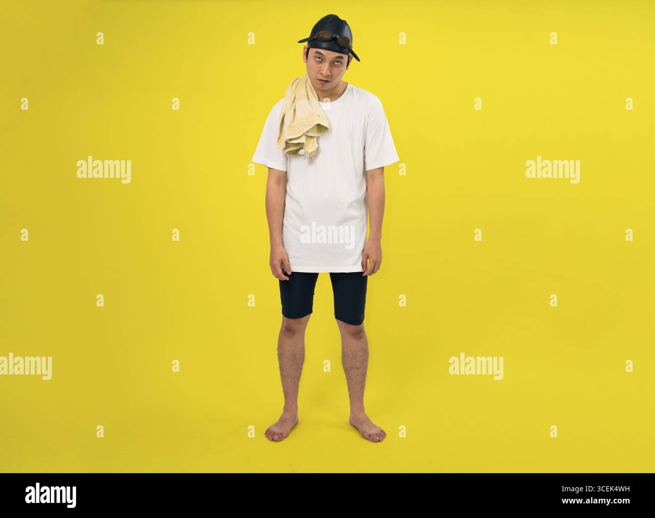 Un uomo vestito con una t-shirt bianca, pantaloncini neri e berretto da nuoto si staglia su uno sfondo giallo brillante. Sembra rilassato e non chalante Foto Stock