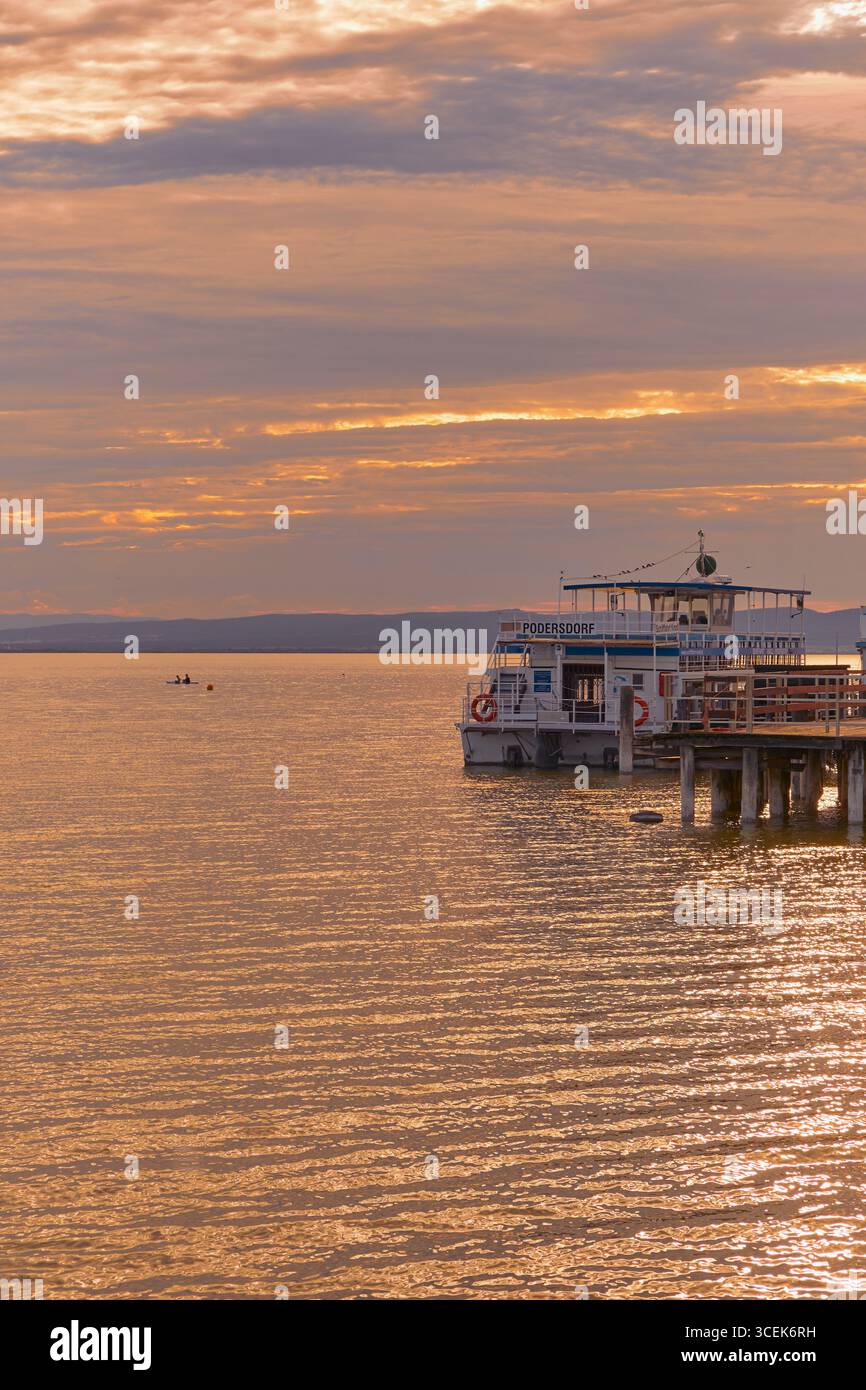 Traghetto passeggeri attraccato presso un molo sul lago di Neusiedl, Austria, durante la luce dorata del tramonto con acqua calma e tranquilla atmosfera estiva. Foto Stock