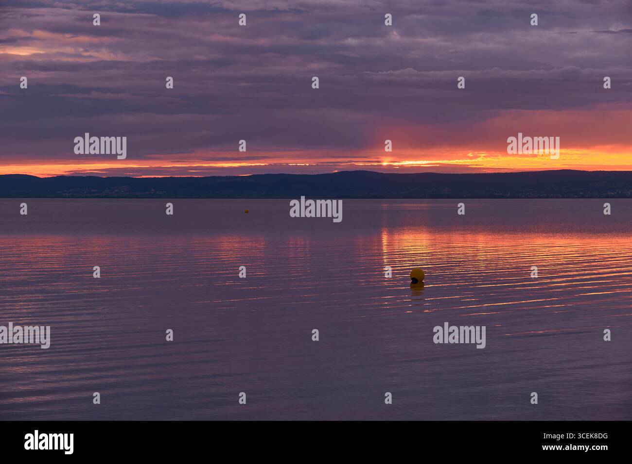 Scena mistica del lago serale con un cielo viola profondo, un brillante bagliore arancione che si riflette sull'acqua calma, sagome di montagna ombrose e una boa solitaria. Foto Stock