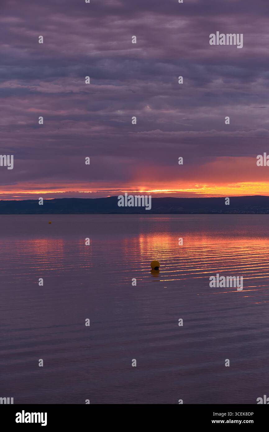 Lago al tramonto con cielo viola, vivace striscia arancione sull'acqua, lontane sagome di montagna e boa nel riflesso luminoso. Foto Stock