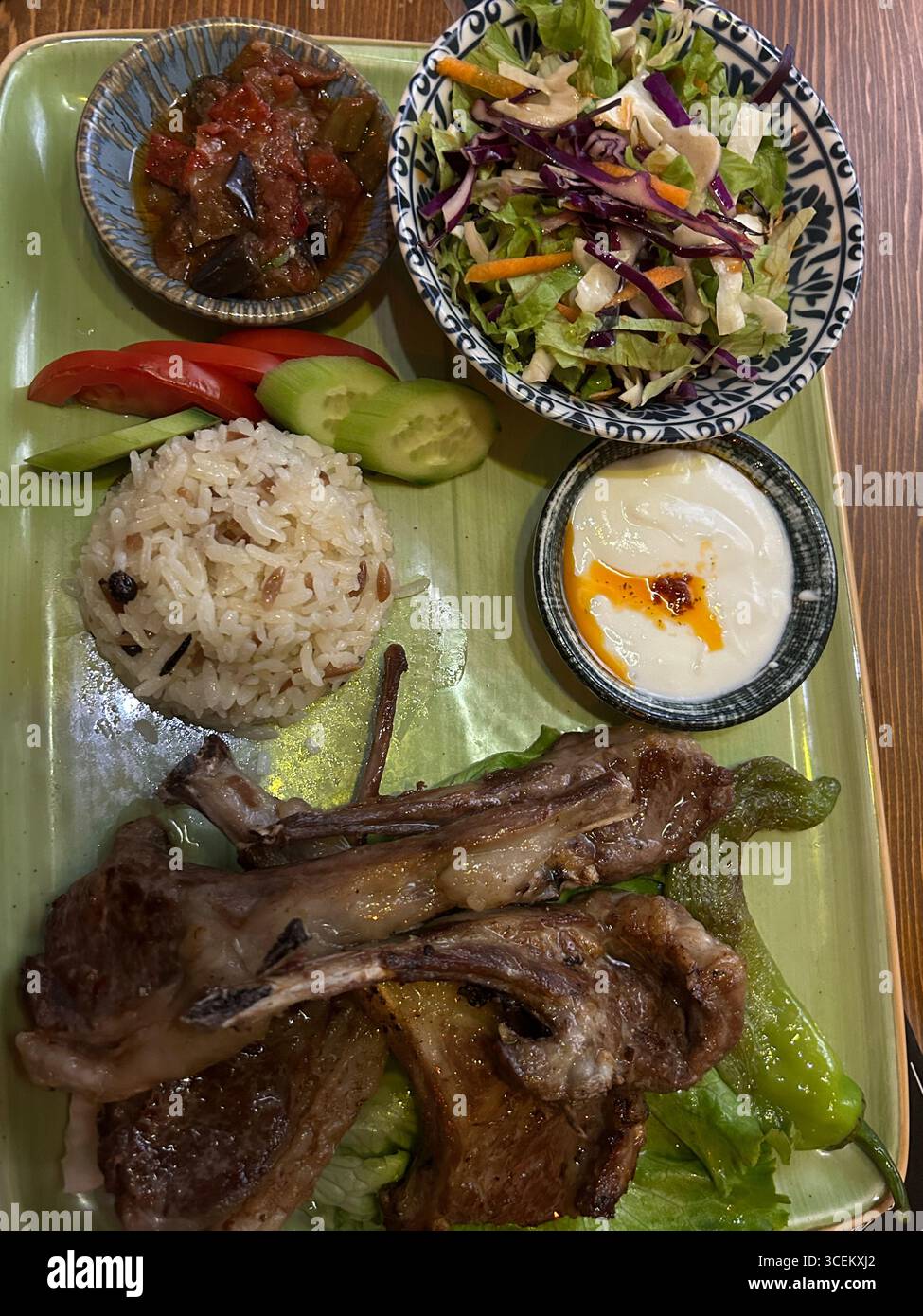 Questa foto mostra un pasto completo di costolette di agnello servito al tavolo di un ristorante di legno. L'impostazione include salse, insalata, riso, yogurt e agnello alla griglia. Il ba Foto Stock