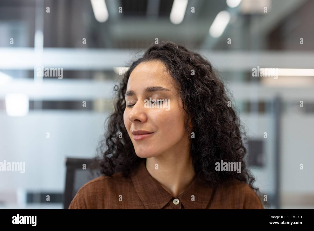 Ritratto di una donna serena sorridente a occhi chiusi, in piedi in un ufficio. Appare in pace, godendo di un momento di calma durante la sua giornata in uno spazio di lavoro professionale. Foto Stock