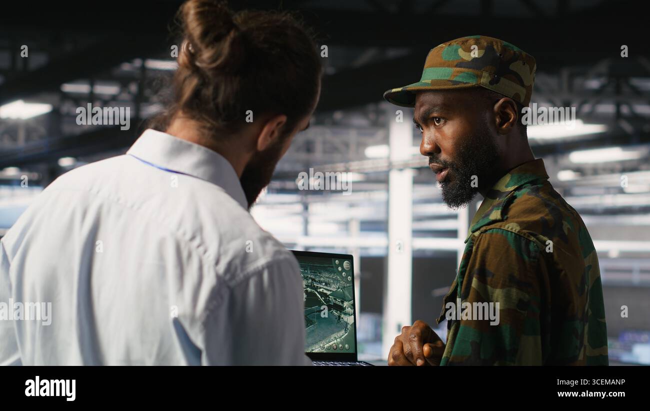 Esperti militari nel data center che esaminano i feed di sorveglianza dei droni sullo schermo dei notebook. Unità dell'esercito nella sala server high-tech che eseguono operazioni di ricognizione sul campo di battaglia utilizzando un notebook Foto Stock