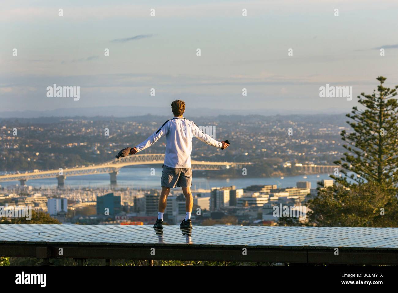 Un giovane si erge con le braccia allungate abbraccia la vista panoramica del ponte del porto e del paesaggio urbano dal Monte Eden, Auckland, nuova Zelanda Foto Stock