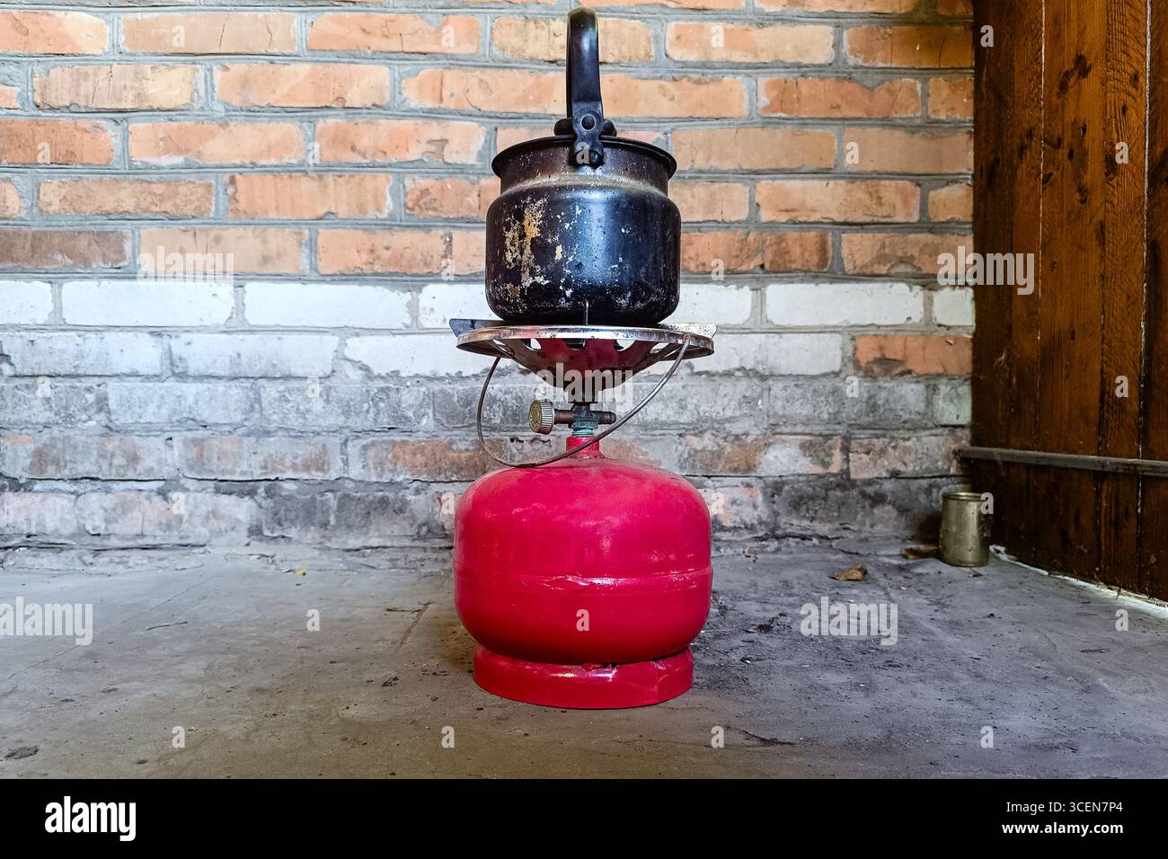 Bollitore nero usurato posizionato su una piccola stufa a gas rossa, appoggiato su una parete rustica in mattoni in un ambiente interno semplice. Foto Stock