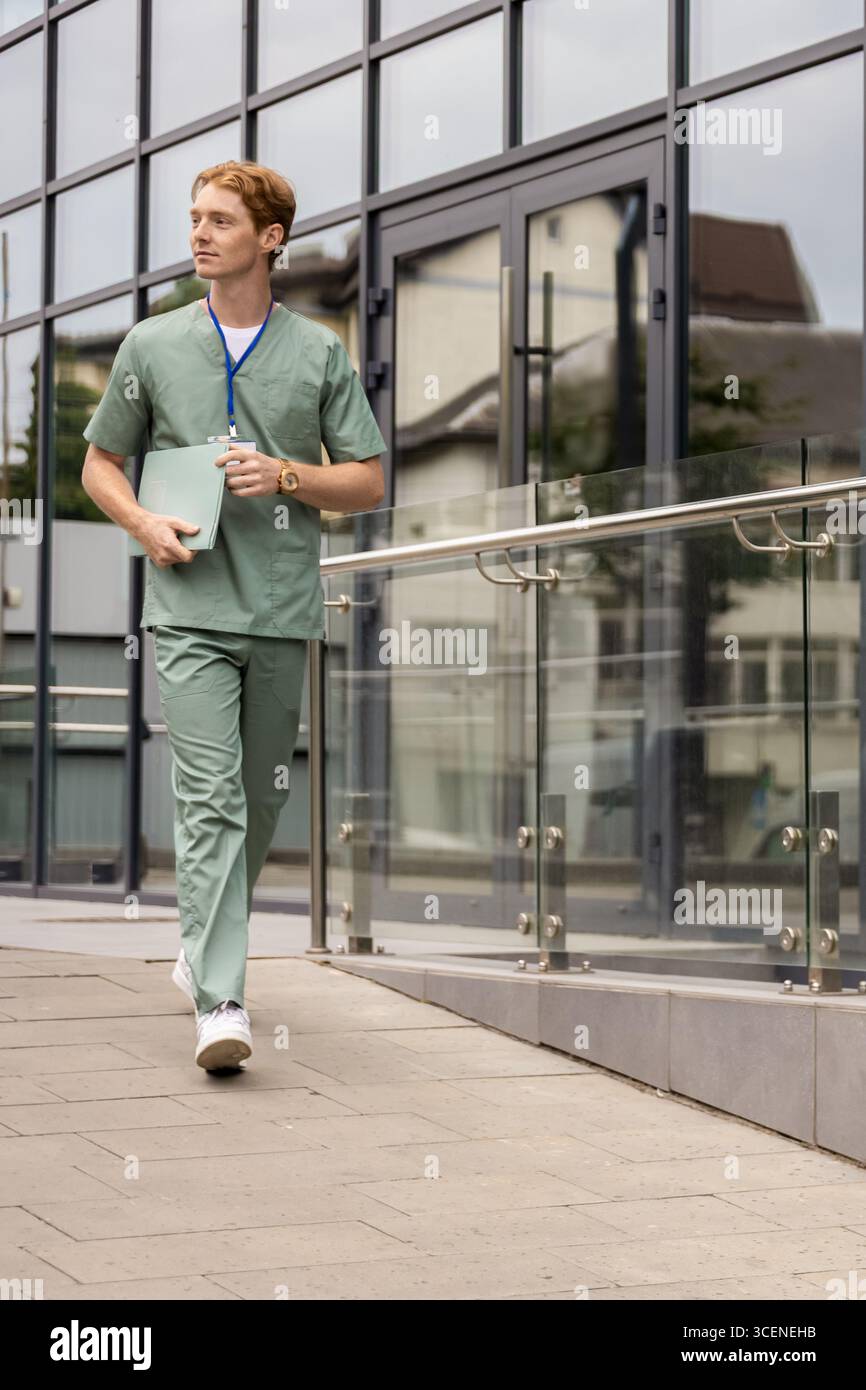 Medico caucasico in uniforme verde che cammina lungo la facciata della clinica con badge e cartellino Foto Stock
