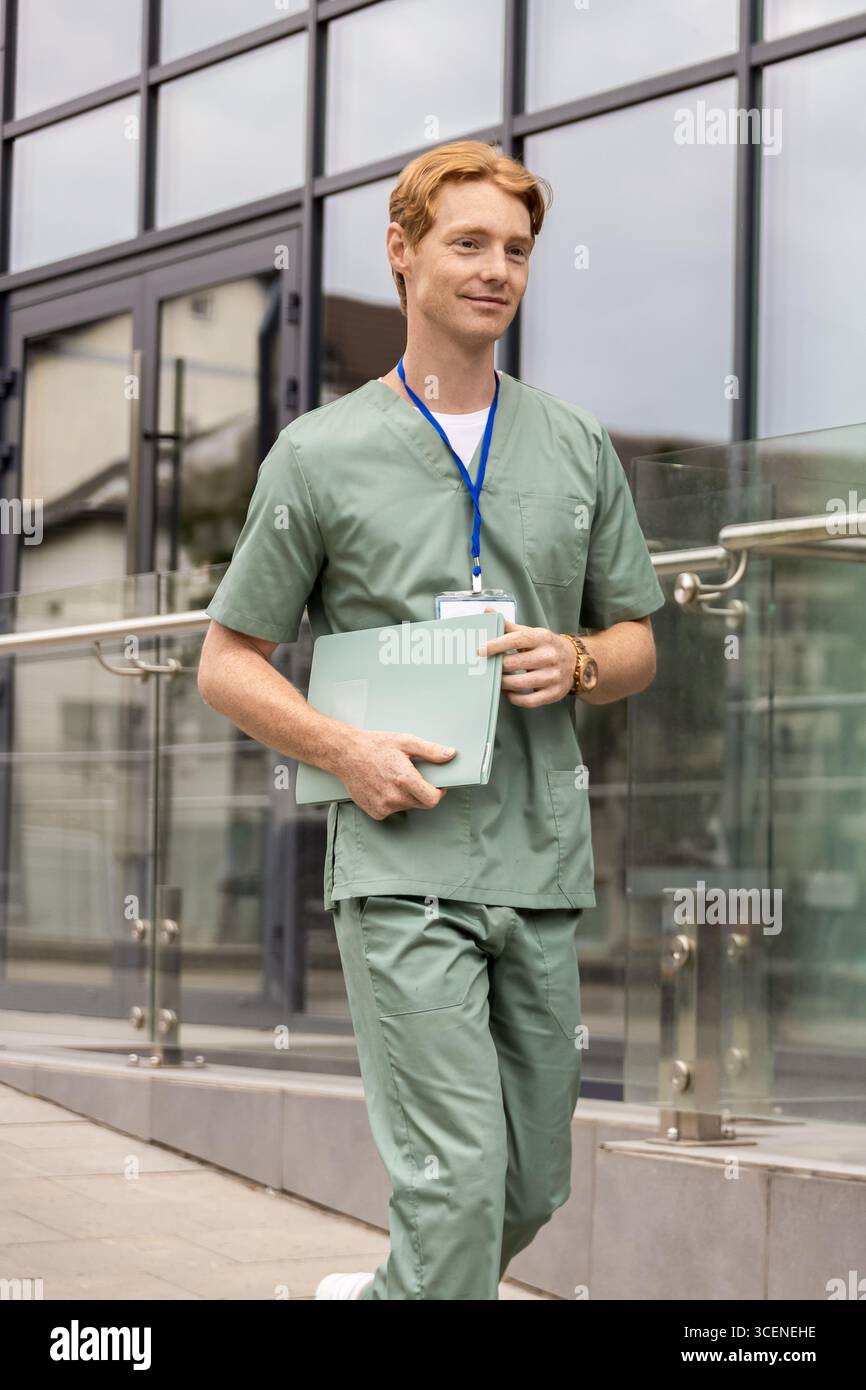 Medico caucasico in uniforme verde che cammina lungo la facciata della clinica con badge e cartellino Foto Stock