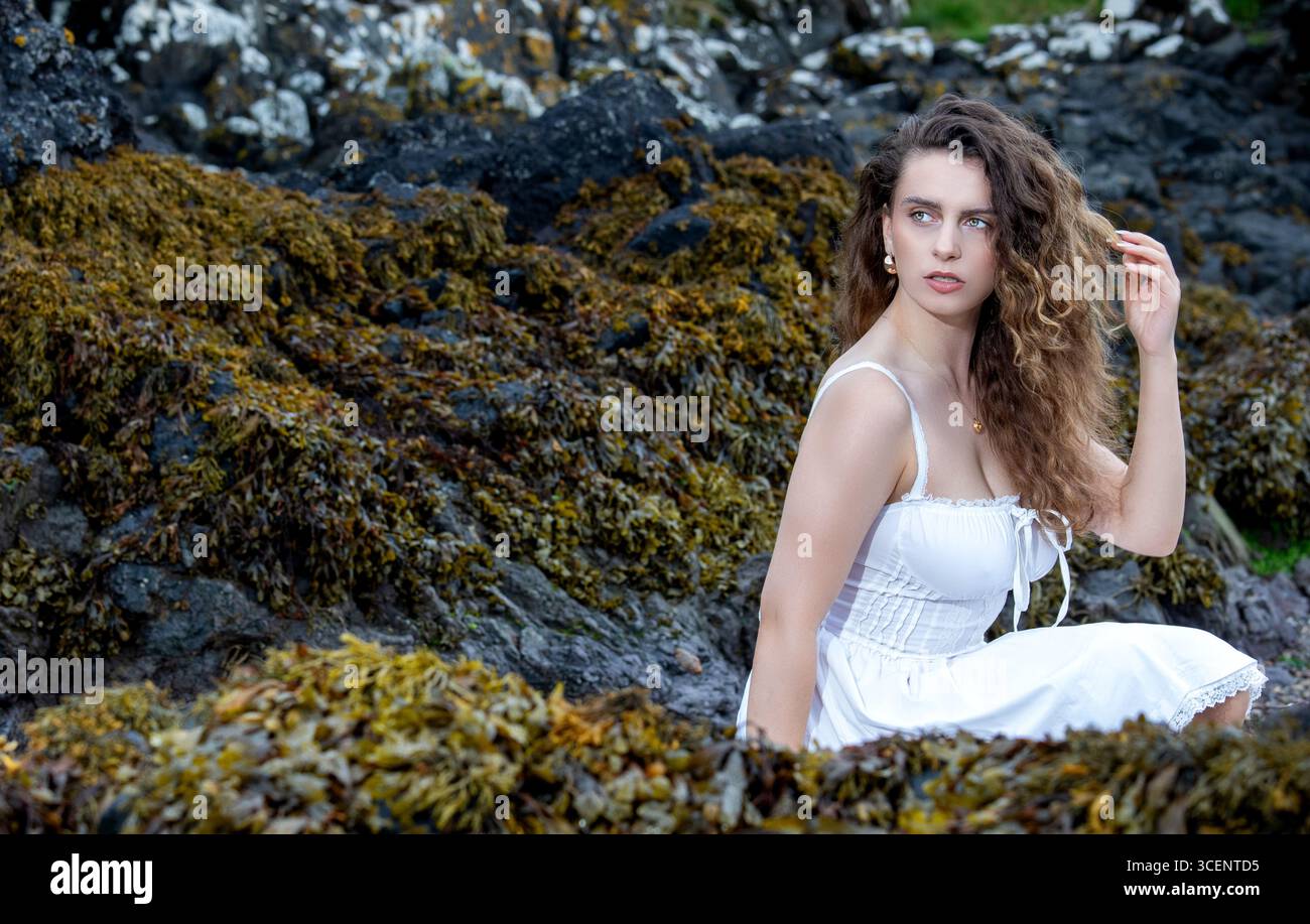 Spiaggia del faro di Tayport a Fife, Scozia, una donna con un abito corto bianco si posa tra le rocce in un mistico servizio a tema fantasy, Regno Unito Foto Stock