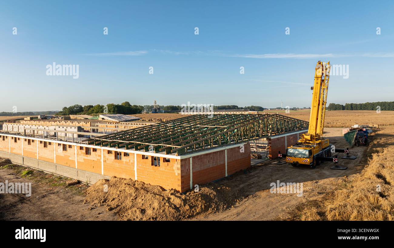 Una gru telescopica mobile posizionata in un cantiere che installa capriate con tetto verde su un grande edificio in mattoni, circondato da terreni agricoli sotto a. Foto Stock