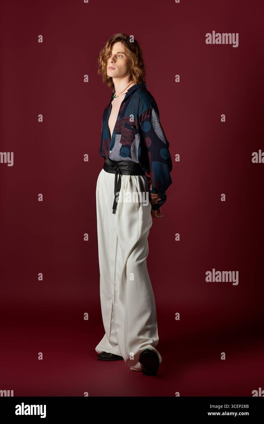 Un giovane uomo trasuda fascino e fiducia in un abbigliamento elegante e femminile sullo sfondo vivace. Foto Stock