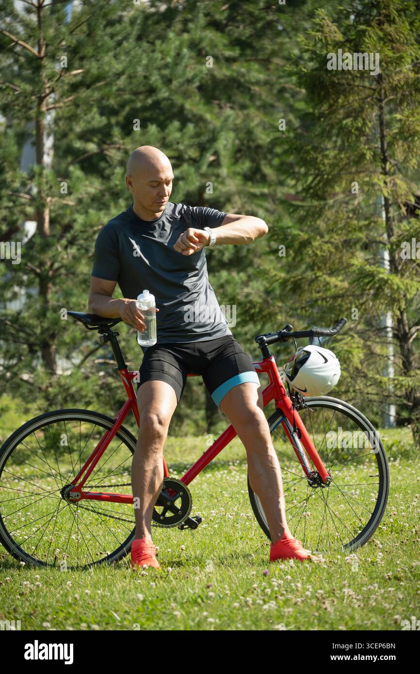 Uomo e ciclista caucasico sportivo con app per il controllo dello smartwatch, progressi di fitness nel parco pubblico. Attività estive all'aperto. Foto Stock