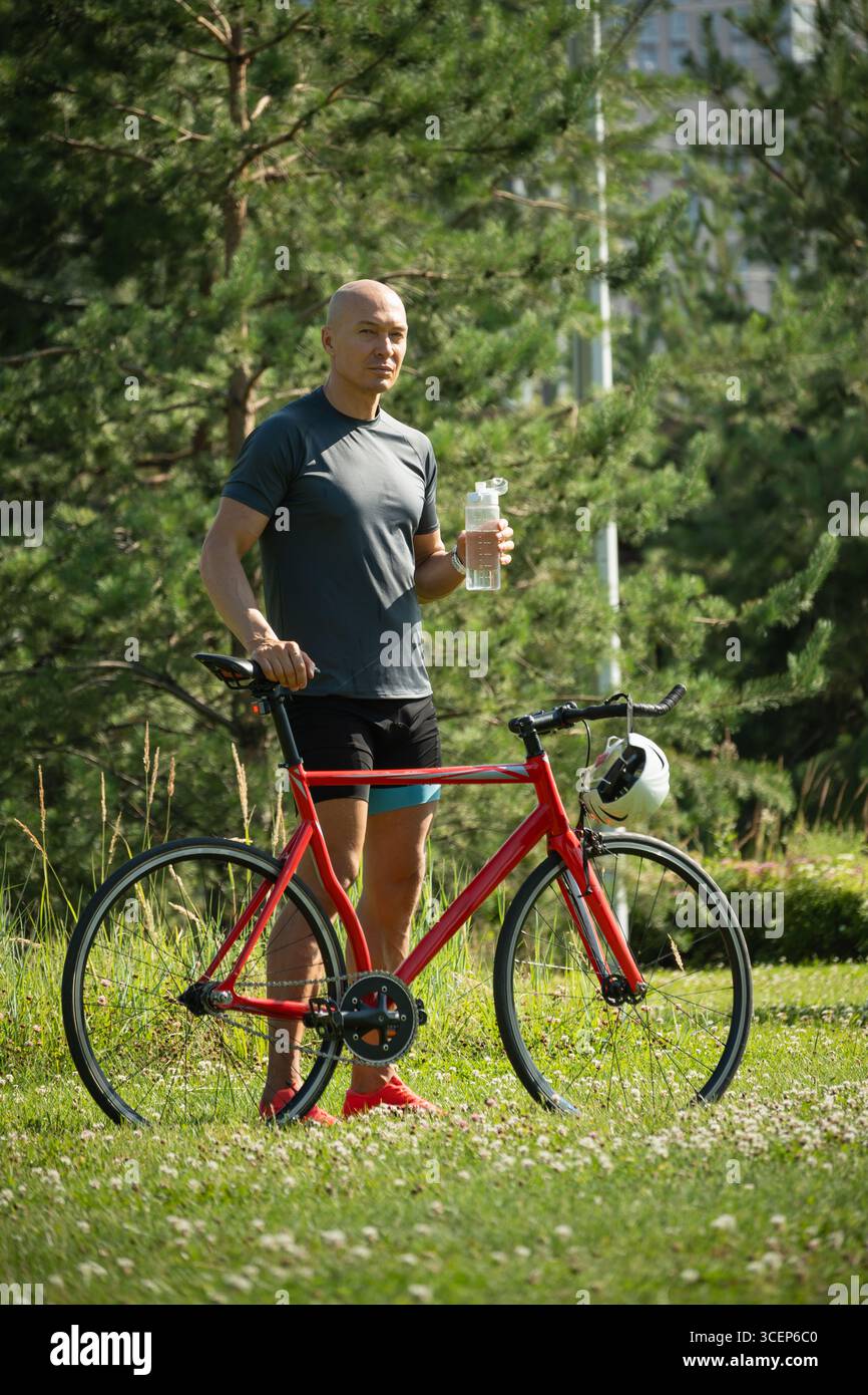 Il ciclista indossa una t-shirt grigia vicino alla bicicletta nel parco pubblico del verde. Attività estive all'aperto. Formato verticale Foto Stock