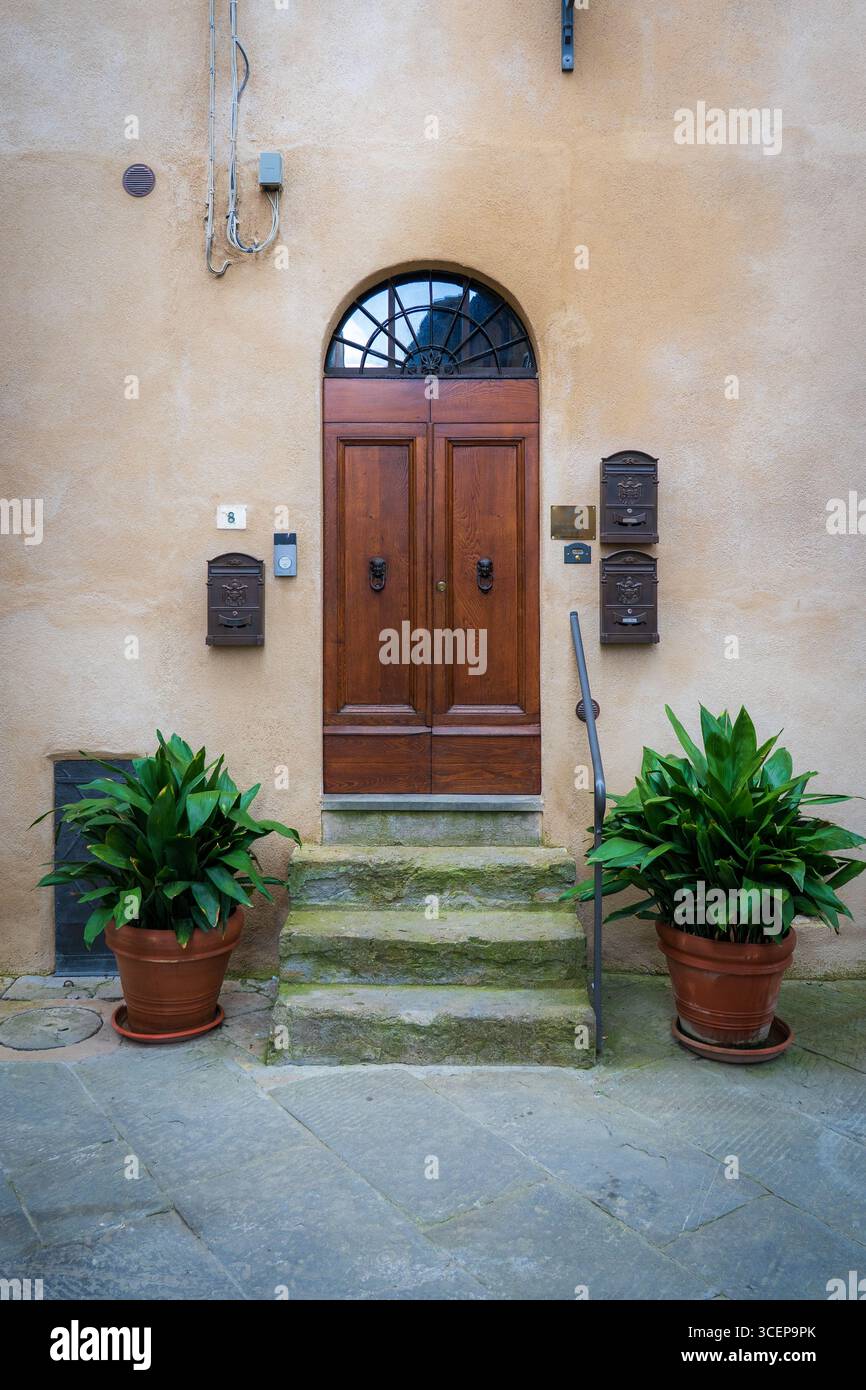Toscana, Italia - 22 aprile 2025: Veduta di una porta ad arco in legno incorniciata da vasi di terracotta di lussureggiante vegetazione, su una parete beige testurizzata, inondata di luce soffusa e diffusa. Foto Stock