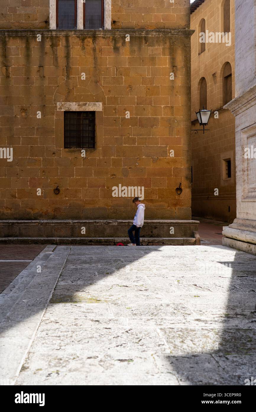 Toscana, Italia - 23 aprile 2025: Veduta di una figura solitaria che attraversa la piazza soleggiata, dove la calda pietra mielata di antichi edifici getta lunghe ombre sul terreno modellato. Foto Stock