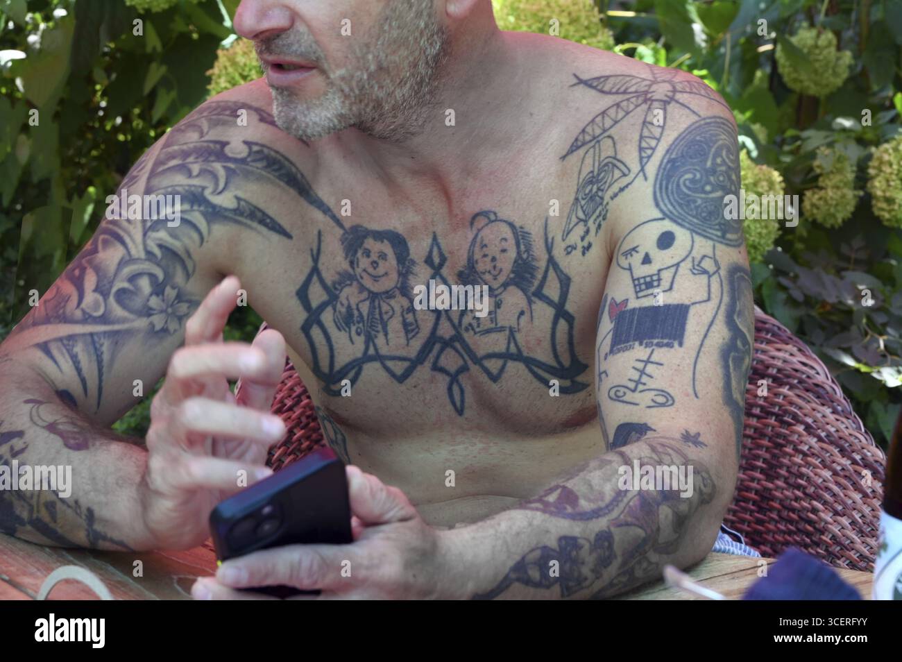 Giovane al telefono cellulare e tatuaggi sulle braccia e sulla parte superiore del corpo, seduto in giardino, Baden-Wuerttemberg, Germania Foto Stock