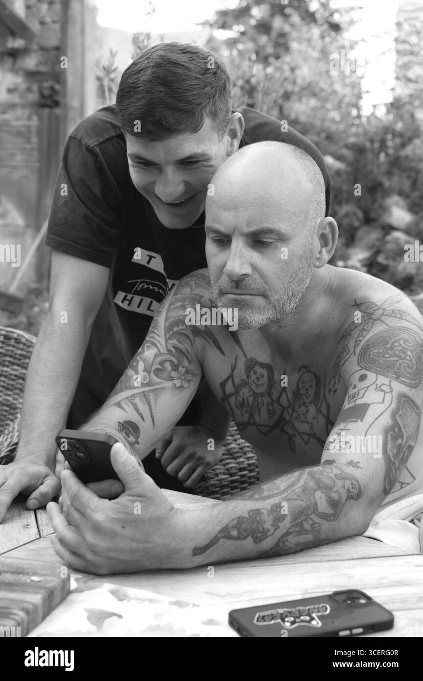 Padre e figlio in giardino al telefono cellulare, padre con tatuaggi sul petto e sulle braccia, Baden-Wuerttemberg, Germania Foto Stock