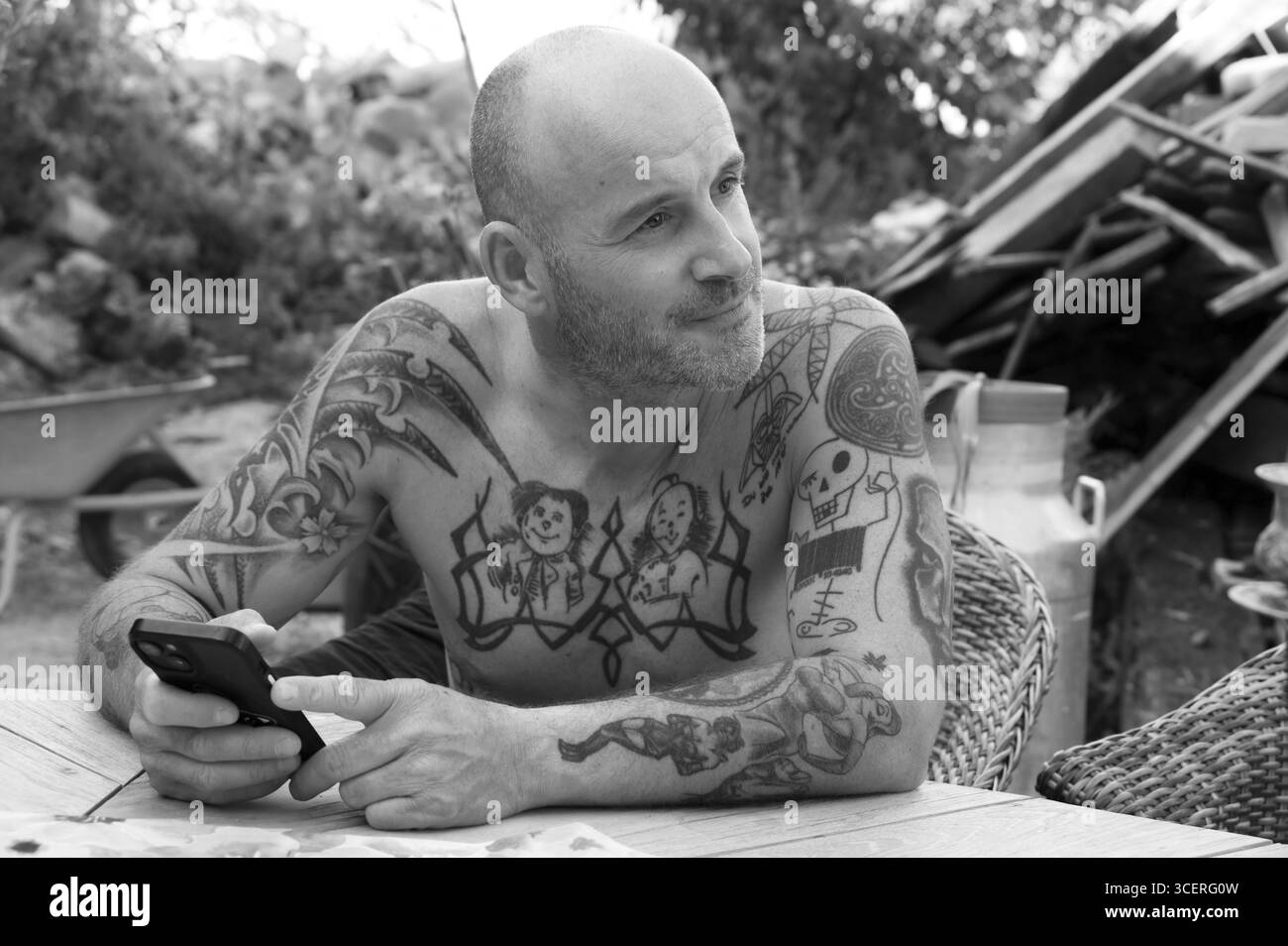 Giovane uomo al telefono cellulare, seduto in giardino con la parte superiore nuda del corpo, tatuaggi sul petto e sulle braccia, bianco e nero, Baden-Wuerttemberg, Germania Foto Stock