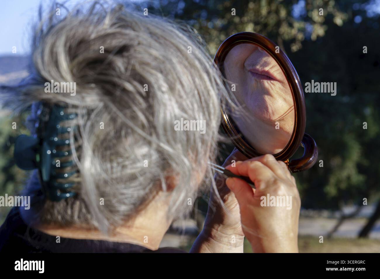 Donna anziana che tiene in mano uno specchio per le mani e raccoglie peli superflui del viso con pinzette alla luce naturale del sole all'aperto Foto Stock
