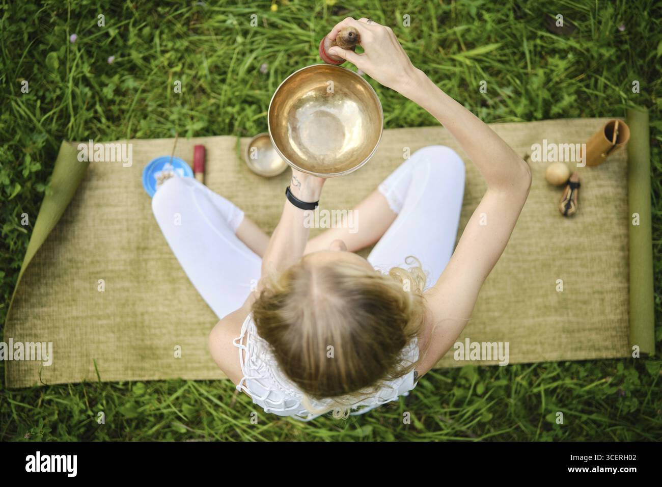 Vista dall'alto di una donna seduta a gambe incrociate su un tappetino, che gioca abilmente una ciotola cantante. Circondato da erba lussureggiante, l'atmosfera tranquilla aumenta Foto Stock