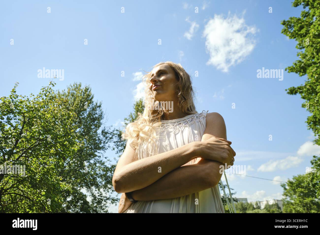 In un parco verde in un giorno luminoso, una donna sta con le braccia incrociate, guardando verso l'alto. La luce del sole mette in risalto i suoi lunghi capelli e il suo abito fluente, creando un Foto Stock