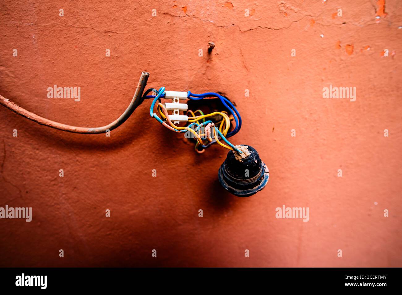 Portalampada esposto con fili colorati aggrovigliati su una parete in terracotta testurizzata. Dettaglio ravvicinato delle infrastrutture obsolete, delle riparazioni stradali e delle util urbane Foto Stock