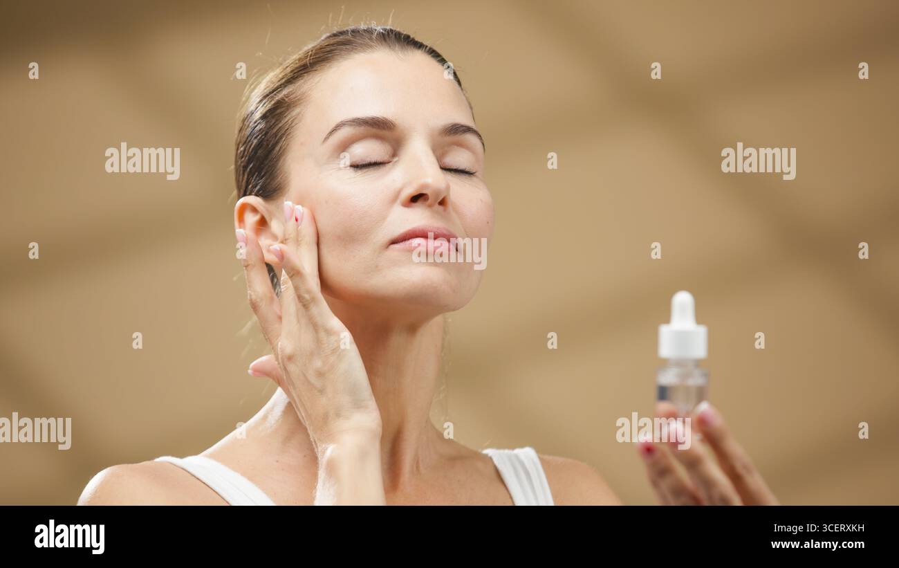 La modella femminile applica delicatamente un siero nutriente sul viso, abbracciando un'atmosfera tranquilla che sottolinea il significato delle routine per la cura della pelle e. Foto Stock