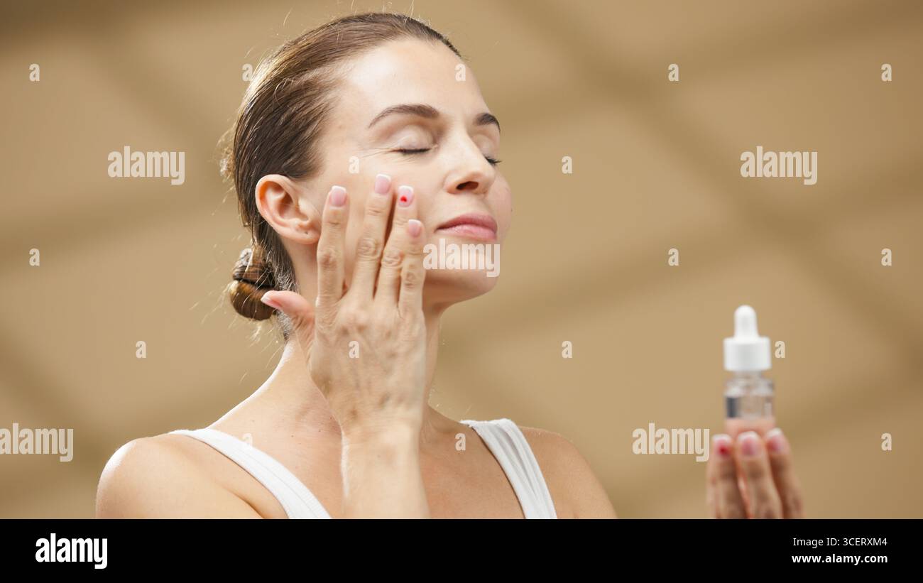 La modella femminile sta applicando il siero al viso con un tocco delicato, mostrando un'espressione serena in un ambiente termale rilassante, sottolineando l'importanza Foto Stock