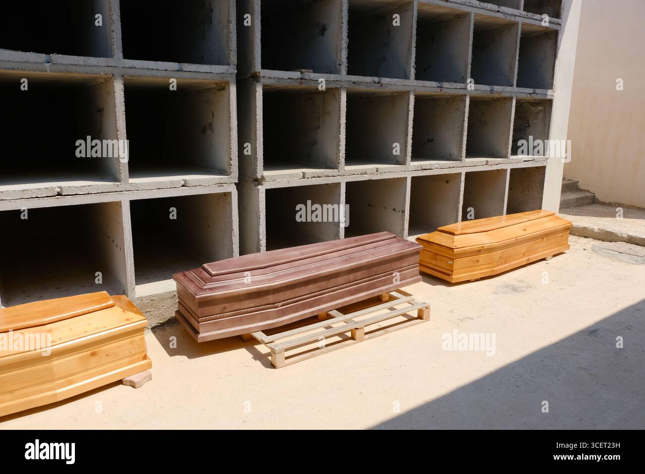 Le bare dei migranti morti nel naufragio del 13 agosto 2025 riposano nel nuovo cimitero di Cala Pisana, Lampedusa. Alcuni recano lettere, probabilmente per l'identificazione. Nella camera mortuaria, le persone si riuniscono in preghiera, mentre i fiori giacciono alla porta. Un tavolo di chirurgo utilizzato per le autopsie si trova nelle vicinanze, sottolineando la tragedia. Le vittime saranno poi trasferite nei cimiteri in Sicilia. Foto Stock