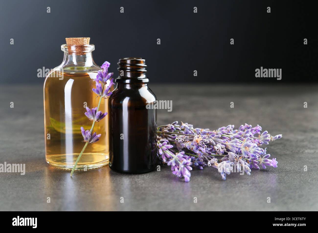 Olio essenziale aromatico in bottiglie e fiori di lavanda su tavola testurizzata grigia su sfondo scuro, primo piano Foto Stock