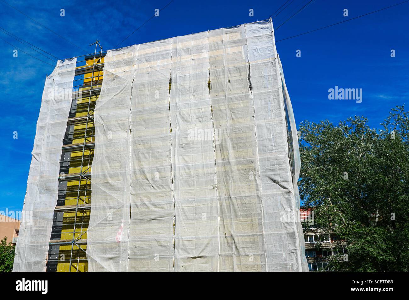 Facciata dell'edificio di appartamenti sottoposta a isolamento termico, coperta da impalcature e rete protettiva contro un cielo azzurro Foto Stock