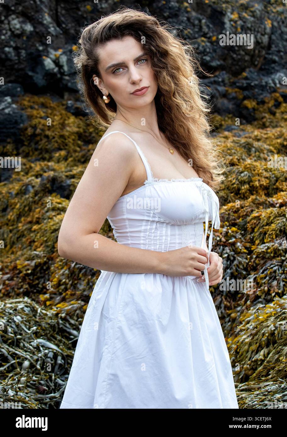 Spiaggia del faro di Tayport a Fife, Scozia. Una donna alla moda che indossa un abito corto bianco si posa vicino alle rocce durante un servizio fotografico a tema fantasy, Regno Unito Foto Stock